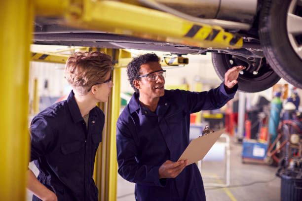 istock Male Tutor With Student Looking Underneath Car On Hydraulic Ramp On Auto Mechanic Course 1218972135