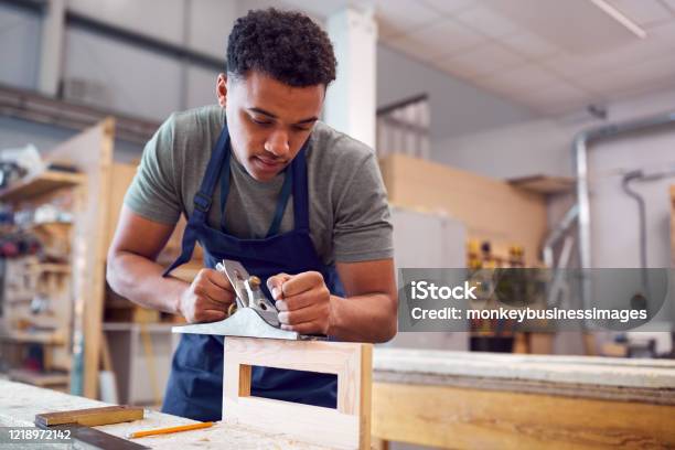 Male Student Studying For Carpentry Apprenticeship At College Using Wood Plane Stock Photo - Download Image Now