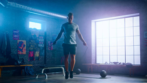 esercizi maschili di giovani atletici con corda da salto in una palestra industriale in stile loft. sta facendo il suo intenso programma di allenamento cross fitness. la struttura ha manifesti motivazionali sul muro. - pantaloncini da corsa foto e immagini stock