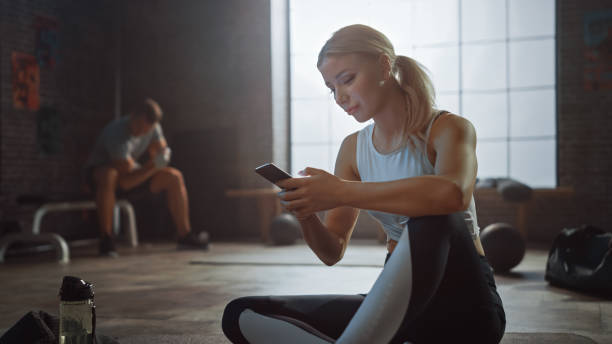 istock Happy and Smiling Beautiful Athletic Young Woman is Using a Smartphone while Sitting on a Floor in a Loft Gym. She's Typing a Message and Smiling. A Man Exercises in the Background. 1218980472