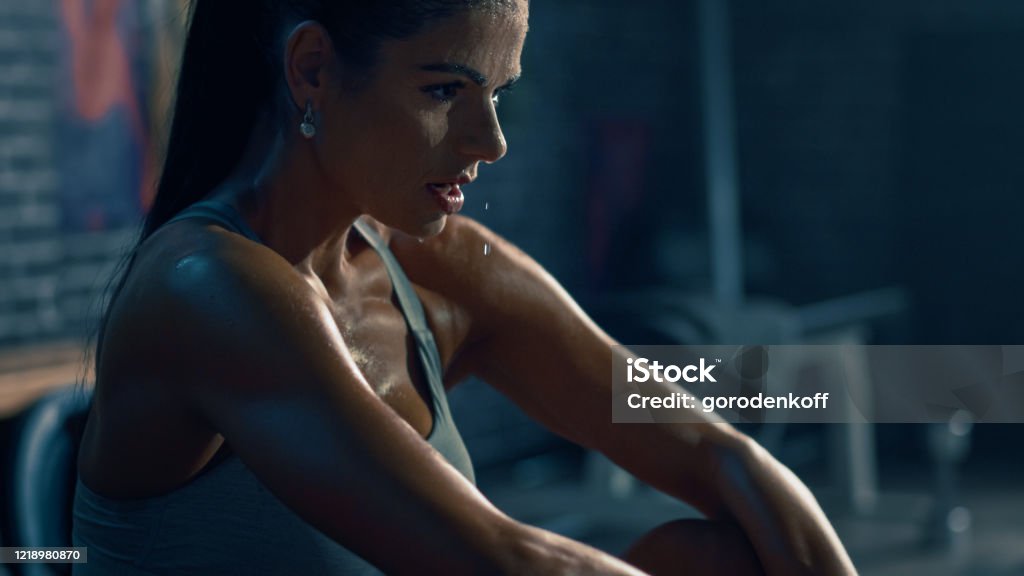 Beautiful Strong Fit Brunette in Sport Top and Shorts in a Loft Industrial Gym with Motivational Posters. She's Catching Her Breath after Intense Fitness Training Workout. Sweat All Over Her Face. Sweat Stock Photo