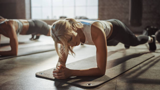 Two Young Fit Atletic Women Hold a Plank Position in Order to Exercise Their Core Strength. They are Exhausted and Struggling with Training. They Workout in a Loft Gym. Two Young Fit Atletic Women Hold a Plank Position in Order to Exercise Their Core Strength. They are Exhausted and Struggling with Training. They Workout in a Loft Gym. bodyweight training stock pictures, royalty-free photos & images