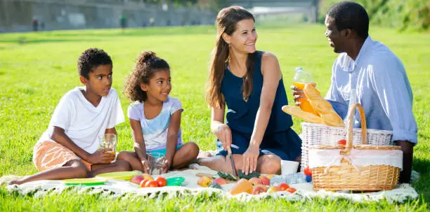 Happy mixed race couple with preteen children gaily spending time at picnic on green lawn