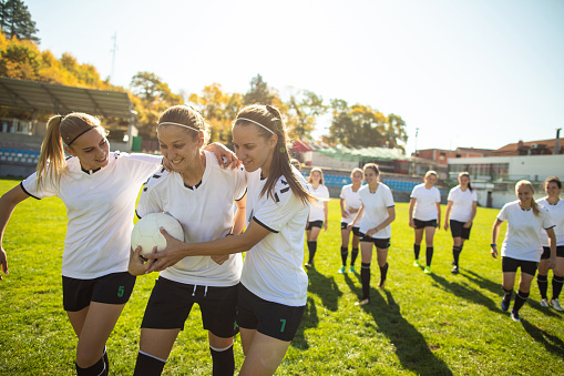 Young female soccer team celebrating a goal, smiling and embracing on soccer field during sunny summer day