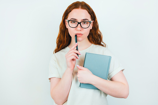 redheaded serious woman in eyeglasses holding pen in one hand near face and planner in another standing on isolated white backgroung, learning and teaching concept.