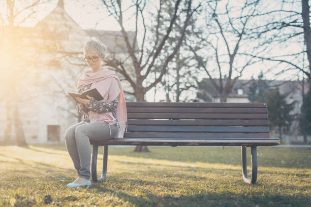 istock Senior woman sitting alone on bench in the park reading a book 1218990664