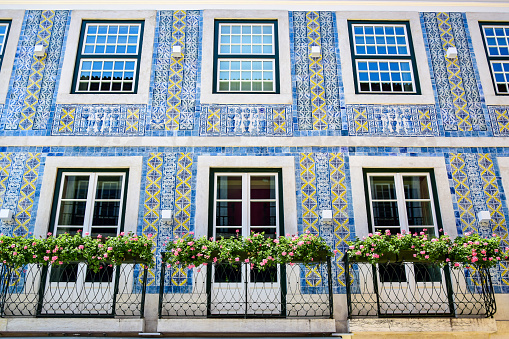 Lisbon, Portugal - 07.23.2019: City house decorated with Portuguese azulejo tiles, blue with white and yellow patterns, white windows and doors, balcony fences decorated with pink flowers.