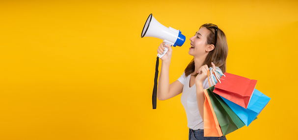 Speaking Loud noise announce of young asian woman with megaphone promotional advertising and shopping paper bag in white t-shirt smiling emotion on yellow background isolated studio with copy space.