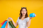 Portrait happy young asian woman holding piggybank saving money for shopping positive emotion in white t-shirt, Yellow background isolated studio shot and copy space.