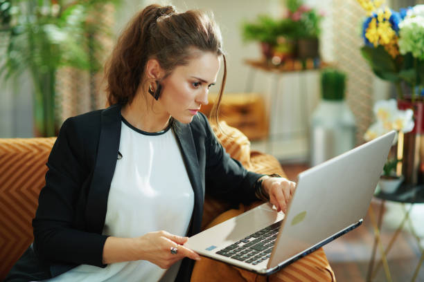 concentrated stylish woman editing while sitting on sofa concentrated stylish housewife in white blouse and black jacket in the modern living room in sunny day editing on a laptop while sitting on sofa. typing on keyboard data breach photos stock pictures, royalty-free photos & images
