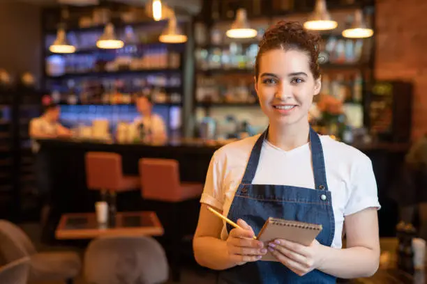 Young smiling waitress in workwear standing in front of camera in luxurious restaurant and going to writie down order of client