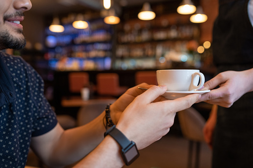 Happy young man taking cup of tea or coffee from hands of waitress on background of bar counter in restaurant or cafe