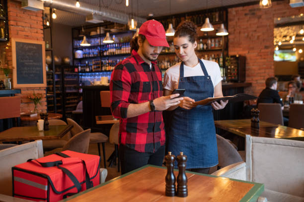 young courier with smartphone and waitress with menu checking online orders - restaurant food food and drink industry food service occupation imagens e fotografias de stock