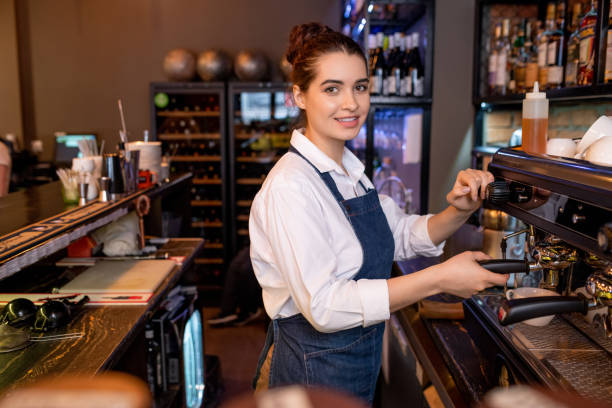 istock Young friendly waitress in workwear preparing cappuccino by coffee machine 1218997196