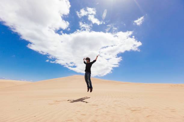 istock Panoramic view of man jumping on a desert dune with blue sky and clouds. 1218998077