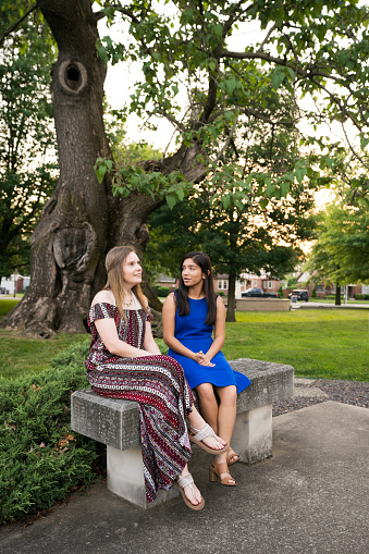 Two high school senior teenage girls sit on a concrete bench under a giant oak tree on a spring day