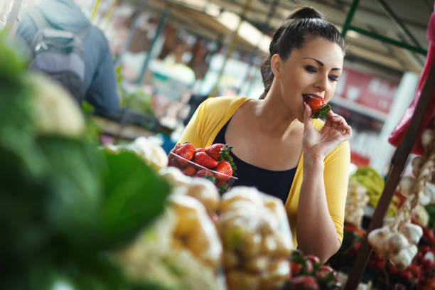 молодая женщина дегустации клубники на продовольственном рынке. - fruit flavor стоковые фото и изображения