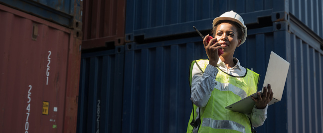 Black foreman woman worker working checking at Container cargo harbor holding laptop computer and walkie-talkie to loading containers. African dock female staff business Logistics import export shipping concept.