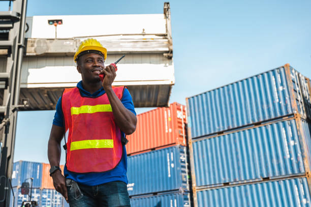 schwarzvorarbeiter steuern den kran und gabelstapler am container frachthafen zum verladen von containern. afrikanische dock männliche mitarbeiter mit radio walkie-talkie für logistik import export versandkonzept. - talkie stock-fotos und bilder