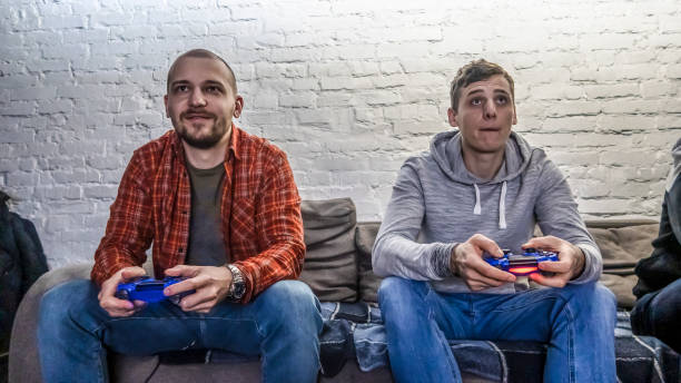 istock Two men playing video game in front of a white brick wall. Hands holding a console controller. Guys sit on the sofa, front view 1219006162