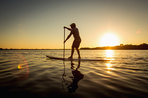 Silhouette of a man on stand up paddle board. Stand up paddler at sunset, paddle board sport. A tourist practicing a water sport during his time off.