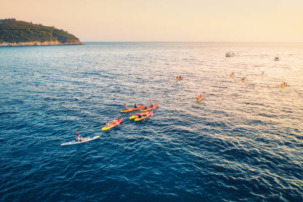 vue aérienne des personnes dans des kayaks dans la mer au coucher du soleil en été. vue supérieure du canot flottant. paysage avec l’eau claire, la montagne, le ciel orange et les bateaux. sport et loisirs. mode de vie et voyages - morning croatia blue sea photos et images de collection