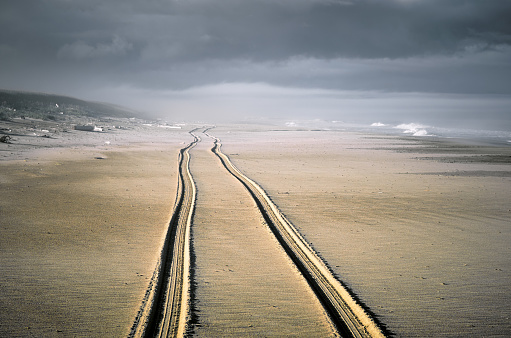 Trail of two wheels of car drove along the wide beach. Coastline sea and dark cloudy sky