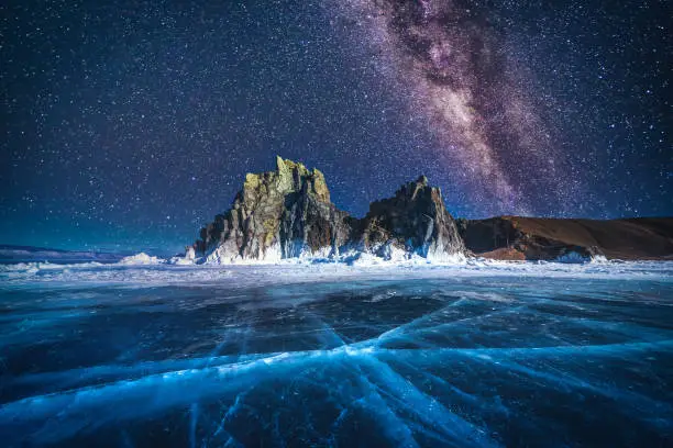 Landscape of Shamanka rock and milky way on sky with natural breaking ice in frozen water on Lake Baikal, Siberia, Russia.