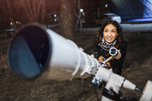 Woman at night looks at the starry sky in a large professional telescope and smiles