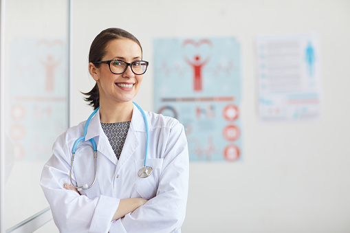 Smiling female doctor in eyeglasses and in white coat working at hospital she standing at her office