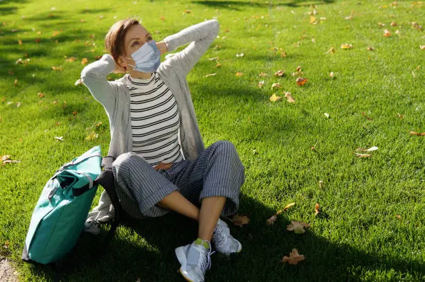 Beautiful woman in protective mask sitting in lonely park on green grass
