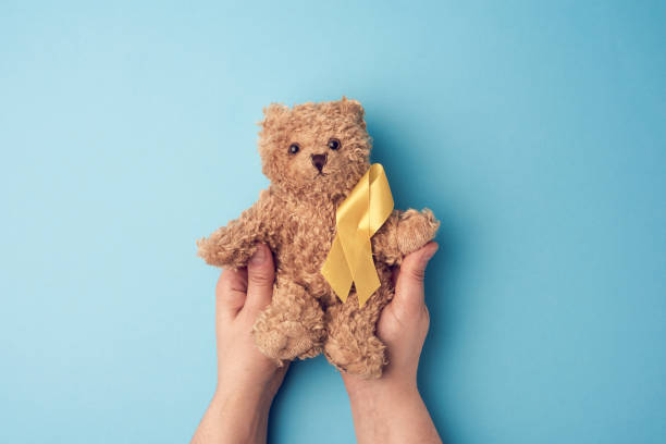 female hands hold a small teddy bear with a yellow ribbon folded in a loop on a blue background female hands hold a small teddy bear with a yellow ribbon folded in a loop on a blue background. concept of the fight against childhood cancer. problem of suicides behavior teddy bear doll old stock pictures, royalty-free photos & images