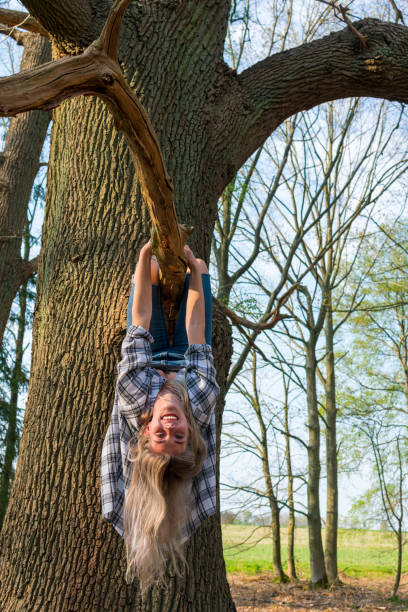 istock Young woman laughingly hangs upside down on a branch of a thick tree 1219050331