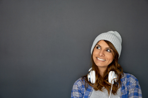 Portrait of a thoughtful hipster woman smiling against the wall at the office with headphones round her neck