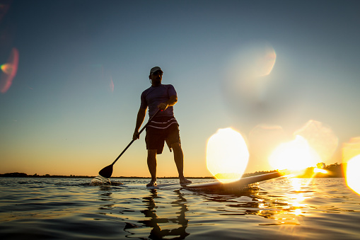 Silhouette of a man on stand up paddle board with water drops in the lens. Stand up paddler at sunset, paddle board sport. A tourist practicing a water sport during his vacations.