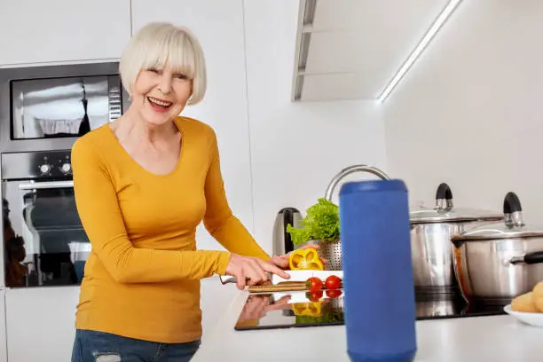 Photo of Retirement. Senior woman standing at kitchen cooking dinner asking to search recipe with wireless smart speaker smiling cheerful