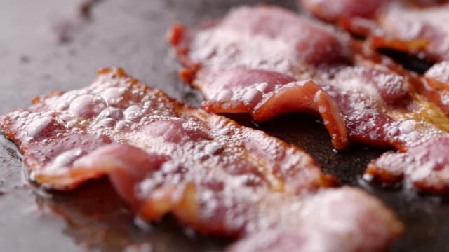 Camera follows cooking bacon on grill. Bacon dolly in slow motion.