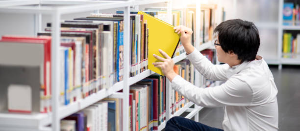 図書館で本を選ぶアジア人男性大学生 - bookshelf book reference book choosing ストックフォトと画像