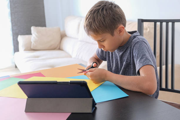 istock Paper crafts for kids. Child cutting colored paper with scissors. Boy looking video tutorial and making paper craft project at home 1219111408