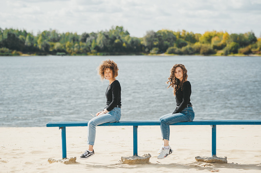 Two brunette girls in jeans are sitting on the bench on the sandy beach of the river. Side view