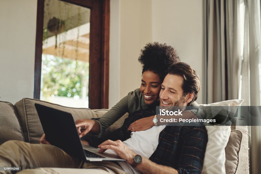 Have a look at that hunny Shot of a happy young couple using a laptop while relaxing on a couch in their living room at home Couple - Relationship Stock Photo