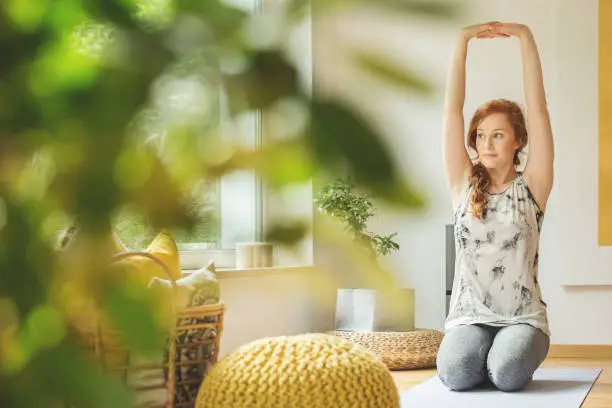 Young woman with outstreched hands-up sitting on yoga mat in room with yellow pouf