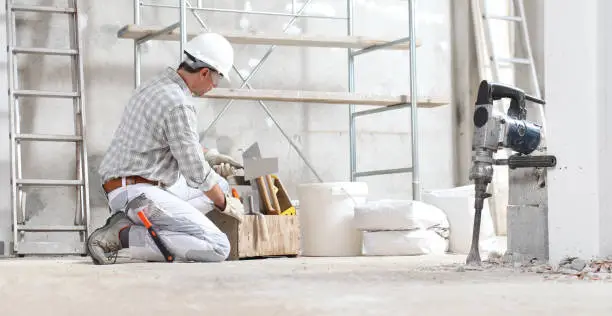 plasterer man construction worker work with tool box wear gloves, hard hat and protection glasses at interior building site with scaffolding. bucket, sacks and jackhammer