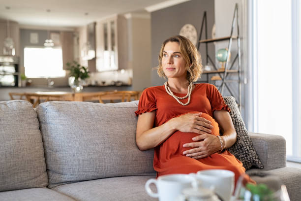 Pregnant woman sitting on couch and thinking Thoughtful middle aged pregnant woman sitting on couch looking away with copy space. Pensive expecting mother relaxing on sofa at home. Beautiful pregnant woman with hands on baby bump thinking about unborn child. smirking stock pictures, royalty-free photos & images