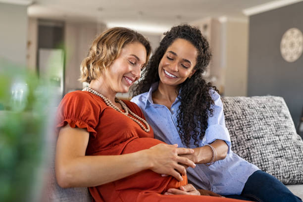 Happy woman hugging her pregnant girlfriend Lovely young lesbian couple planning the future of their baby. Happy friend feeling belly of mid pregnant woman. Happy mixed race woman smiling with hands on expecting mother's baby bump while feeling baby movement; assisted fertilization and in vitro fertilisation concept. Surrogacy stock pictures, royalty-free photos & images