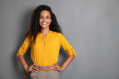 Portrait of young african woman standing with hands on waist and looking at camera. Confident stylish latin girl standing against grey background. Happy young mixed race woman smiling isolated on gray wall with copy space.