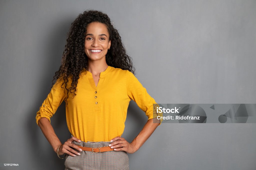 Portrait of beautiful young african woman smiling Portrait of young african woman standing with hands on waist and looking at camera. Confident stylish latin girl standing against grey background. Happy young mixed race woman smiling isolated on gray wall with copy space. Women Stock Photo