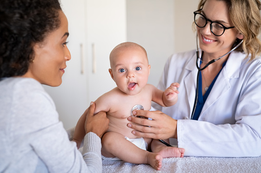 Female smiling doctor examining little baby with stethoscope in clinic. Portrait of a cute little boy checked by doctor with his mother. Pediatrician examining baby boy with a stethoscope.