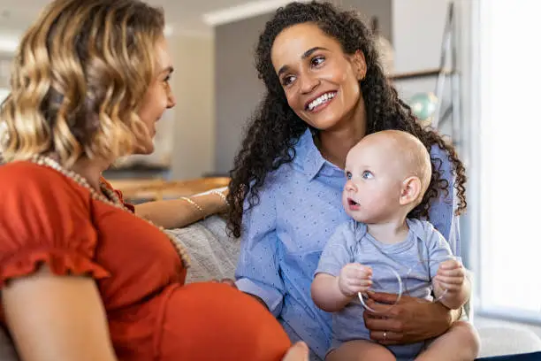 Happy multiethnic friends with child relaxing on couch at home. Mixed race woman with expecting mother talking and playing with baby. Pregnant lesbian mothers sitting on sofa and smiling, assisted fertilization and reproduction concept.