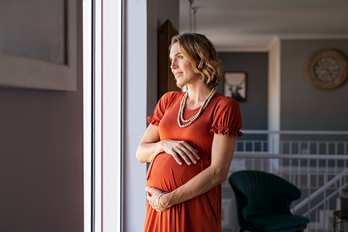 Portrait of mature pregnant woman at home looking outside the window during maternity. Thoughtful pregnant woman standing near window and thinking about the future family. Smiling lady in gestation caressing her baby bump with pensive look.